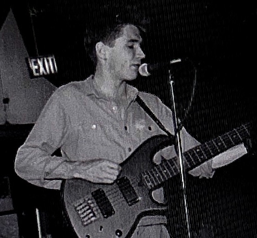 A musician performing on stage, holding an electric guitar and singing into a microphone, with a visible 'EXIT' sign in the background.
