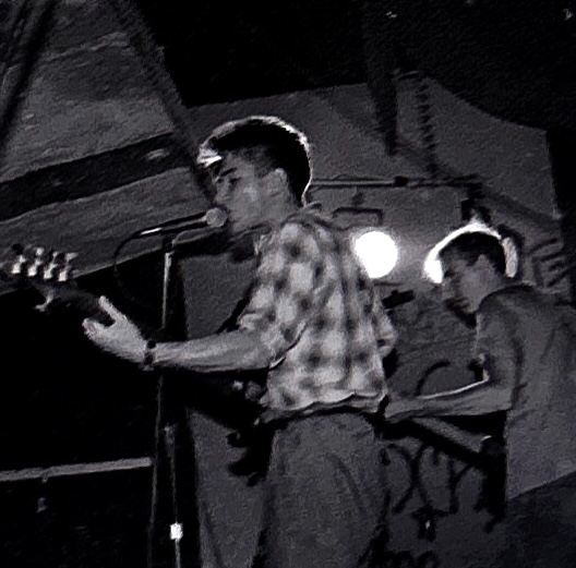 A black and white image of two musicians performing on stage, one singing into a microphone while playing electric guitar, and the other playing guitar in the background.